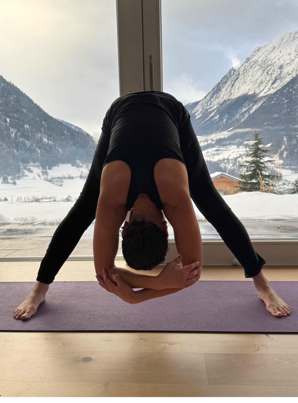 Sarah practising wide-legged forward fold yoga pose with snow-covered Swiss Alps visible through panoramic window in Le Châble, Val de Bagnes